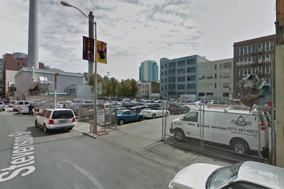 A surface parking lot, full of cars, is located between tall buildings and urban density.